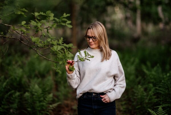 A woman with blonde hair and glasses stands in a lush green forest during the daytime, gently holding a small branch with fresh green leaves. She is wearing a light beige sweater and dark jeans, exuding a calm and thoughtful presence. The surrounding forest is filled with ferns and trees, providing a peaceful and natural atmosphere. The woman appears to be observing the branch closely, as soft light filters through the trees.