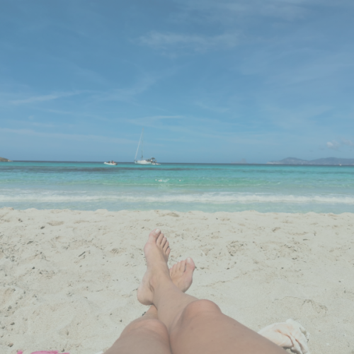 A person resting on a sandy beach with turquoise sea and sailboats in the distance, capturing a peaceful summer holiday moment and deep relaxation.