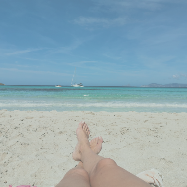 A person resting on a sandy beach with turquoise sea and sailboats in the distance, capturing a peaceful summer holiday moment and deep relaxation.
